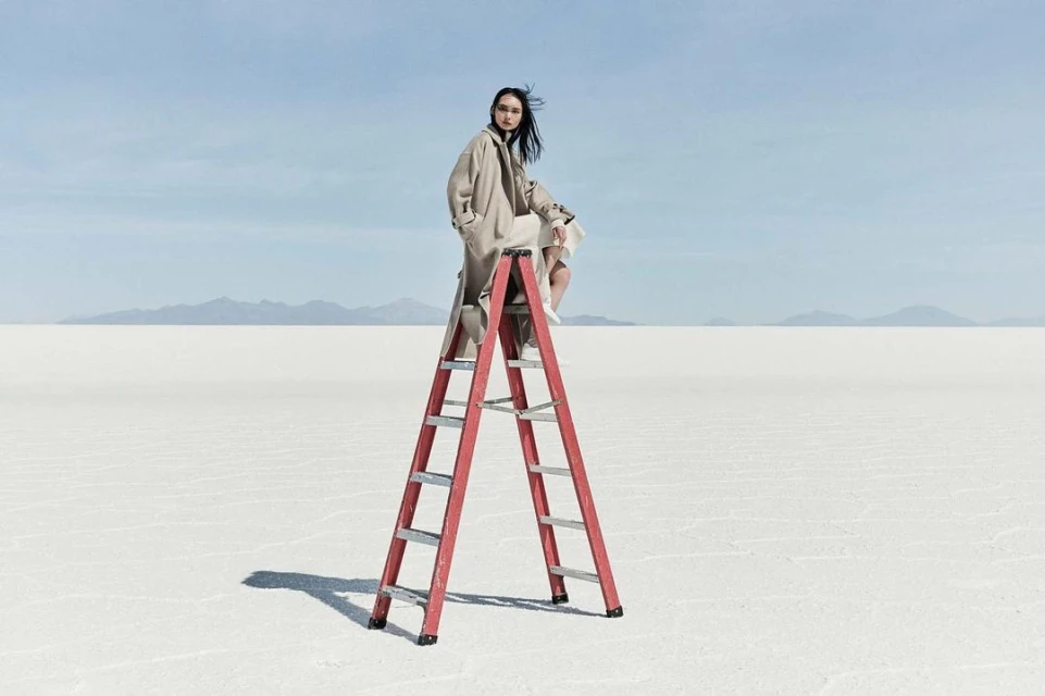 A woman in a beige coat sits on a red ladder in a vast, light desert landscape under a blue sky with distant mountain range.