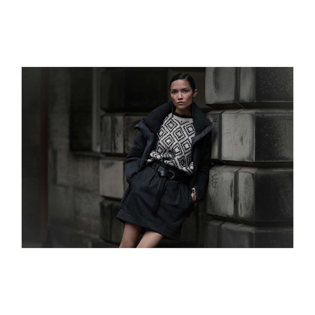 A woman in a sweater and skirt leans against a textured wall outdoors. She's wearing a dark jacket and has her hair pulled back.