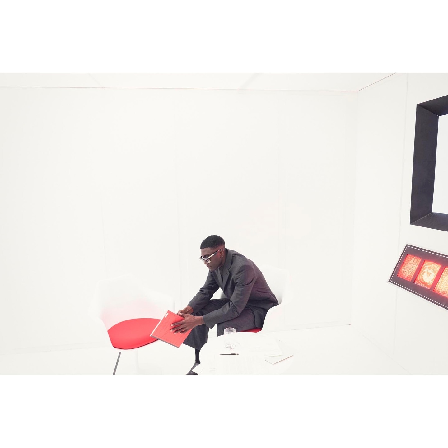 A Black man in a grey suit sits in a white photo studio on a red and white chair, holding a red folder near a white table.