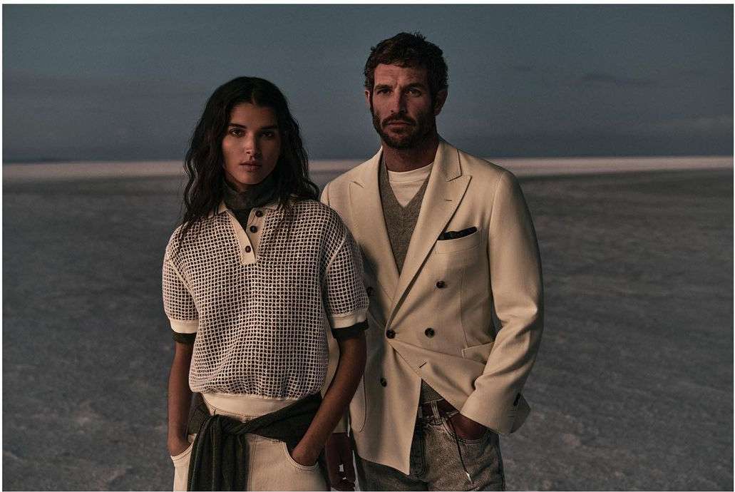 A stylish couple poses outdoors. The woman wears a patterned knit polo, while the man sports a cream-colored blazer in a fashion-forward shot.