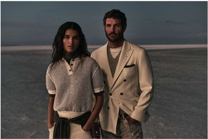 A stylish couple poses outdoors. The woman wears a patterned knit polo, while the man sports a cream-colored blazer in a fashion-forward shot.