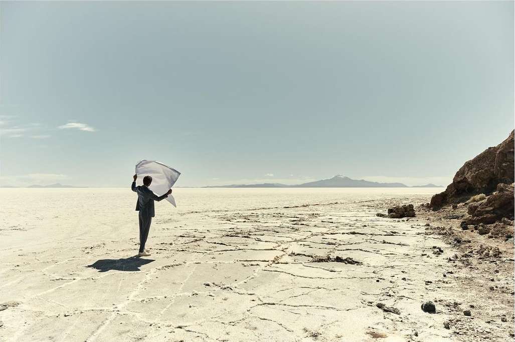 Adult in suit stands on cracked desert holding semi-transparent sheet against bright sky. Mountain range visible in the distance.