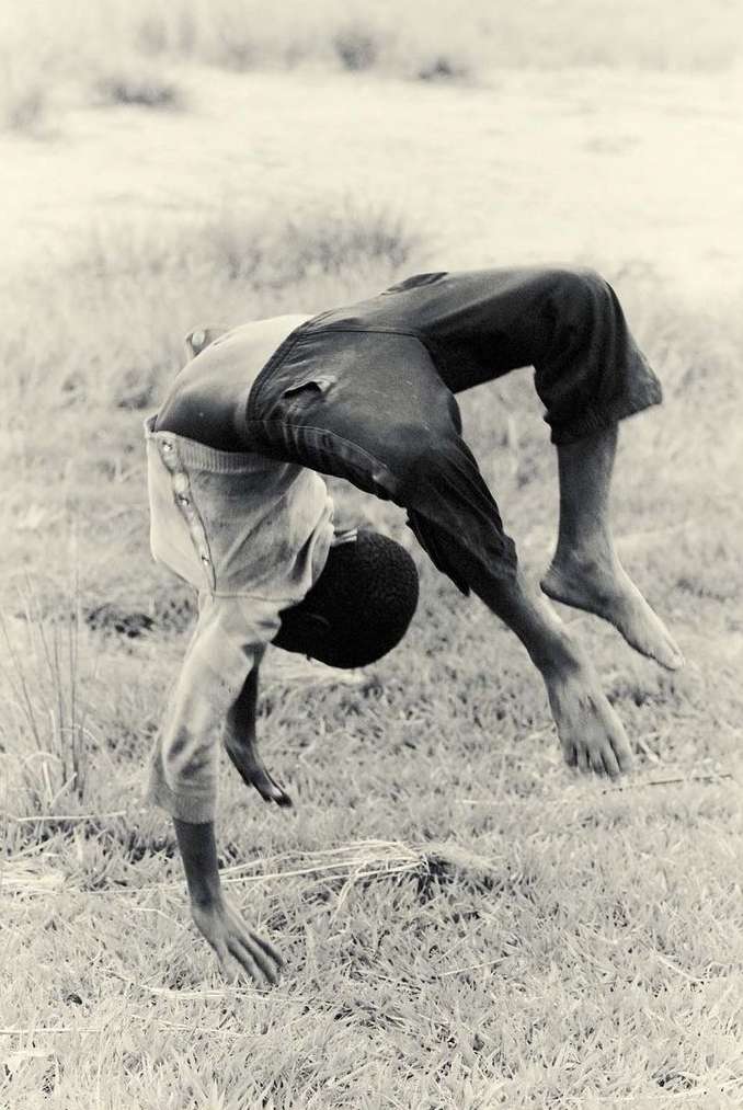 Boy performs a backbend on grassy field, with an out-of-focus background in a monochromatic outdoor photograph.