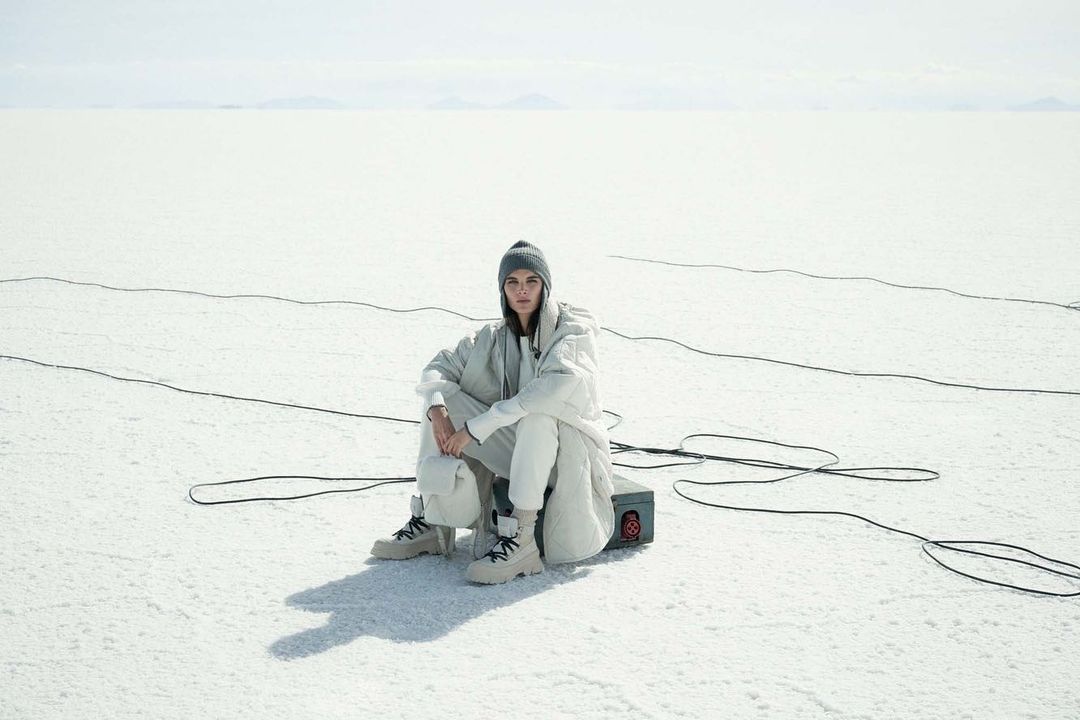 Woman in light-colored clothes sits on a box in a bright, white outdoor setting with tangled cables, wearing a beanie and boots.