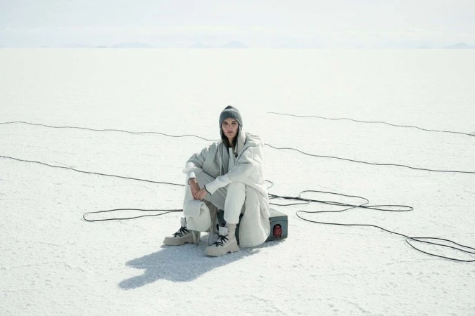 Woman in light-colored clothes sits on a box in a bright, white outdoor setting with tangled cables, wearing a beanie and boots.
