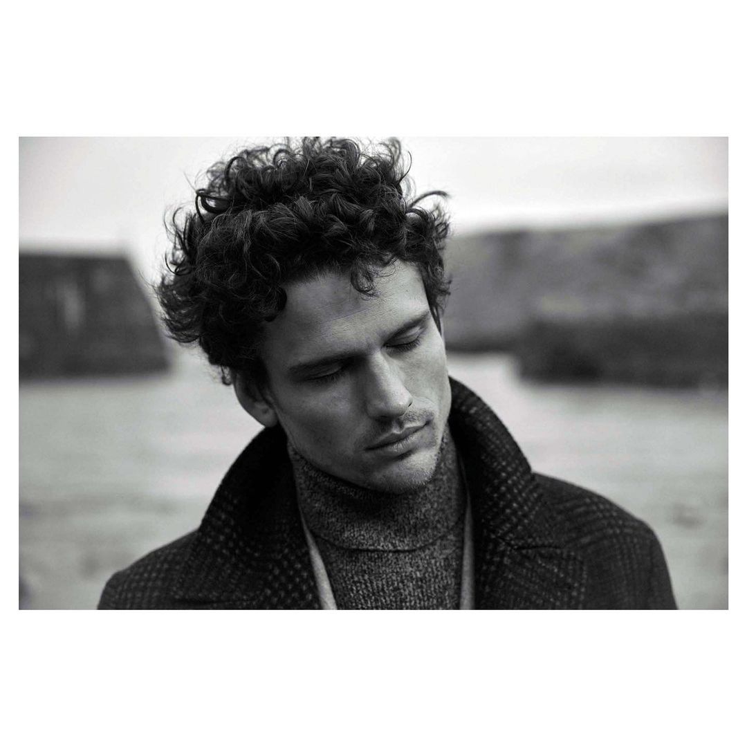 Monochrome portrait of a man with curly hair wearing a turtleneck and textured coat outdoors.