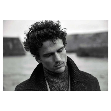 Monochrome portrait of a man with curly hair wearing a turtleneck and textured coat outdoors.