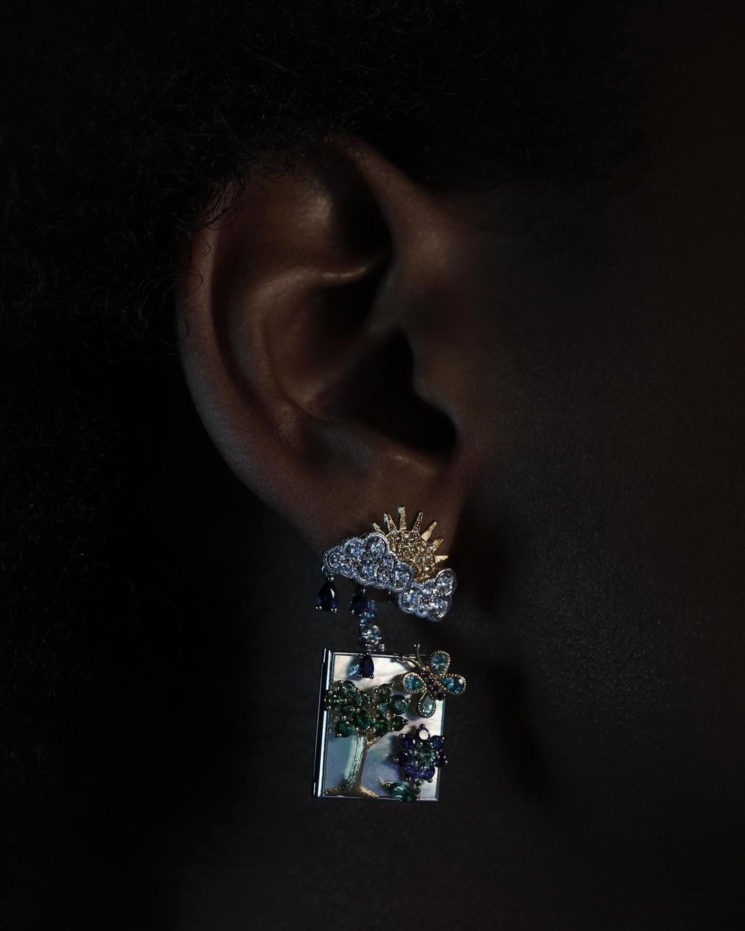 Close-up studio shot of a person's ear wearing an elaborate earring with a cloud, sun, raindrops, and tree design against a dark background.