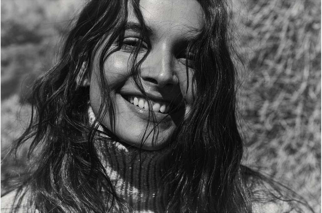 Close-up portrait of a smiling young woman with long, dark hair wearing a turtleneck sweater, photographed outdoors in black and white.