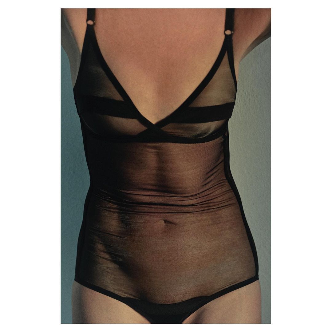 Slender woman in black mesh lingerie bodysuit, indoor setting with soft light. Fashionable and alluring style, enhancing her silhouette.