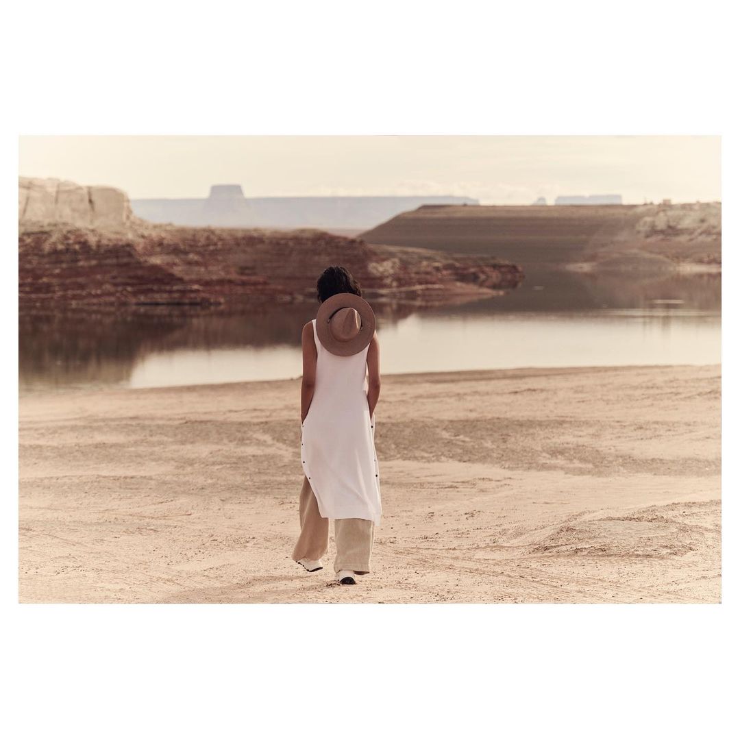 Back view of an adult woman in a white dress and tan hat walking on a sandy beach with a lake and rocky cliffs in the background.