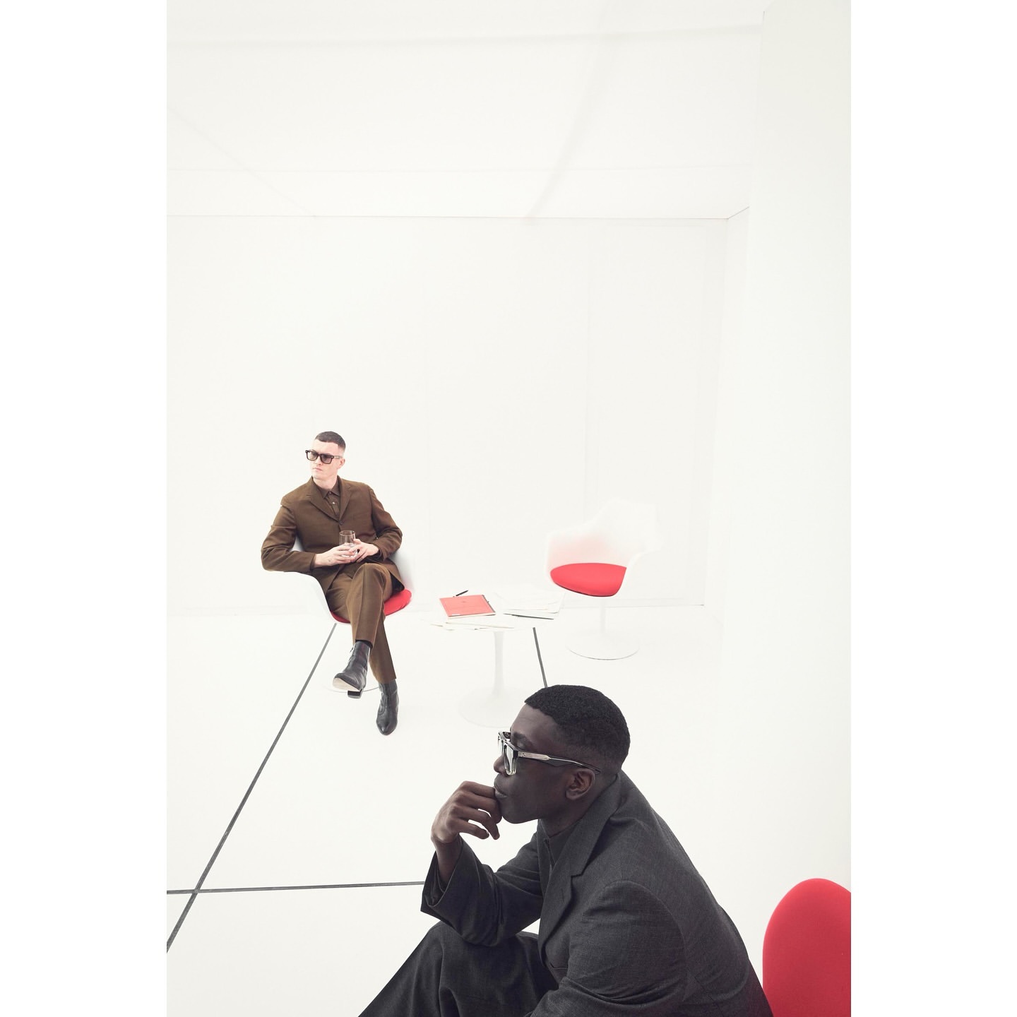 Two men in suits sit on red chairs in a minimalist white room. One man is closer to the camera than the other. 