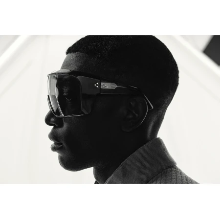 Profile portrait of a Black man wearing oversized black sunglasses and a textured collared shirt against a bright background.