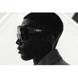 Profile portrait of a Black man wearing oversized black sunglasses and a textured collared shirt against a bright background.