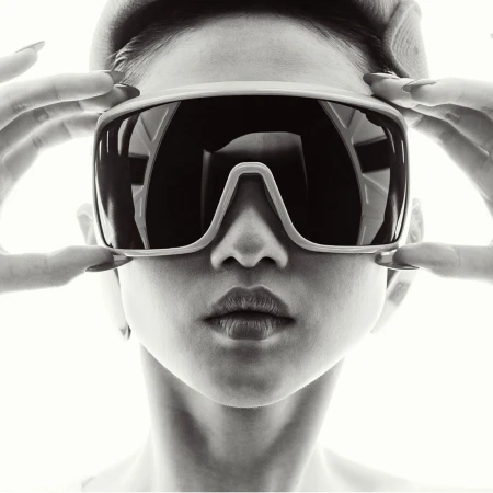 Close-up portrait of a person wearing large visor sunglasses and a brimmed cap, holding the frame with both hands against a bright white background.