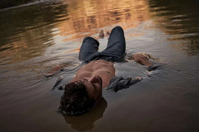 Shirtless man floating on his back in shallow river water wearing dark denim jeans by Alistair Taylor-Young