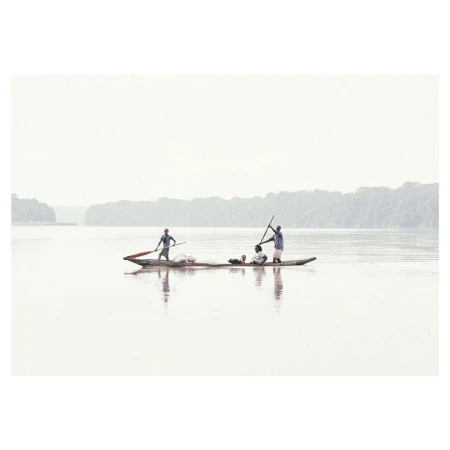People in a long wooden canoe paddling across a wide misty river with distant tree-lined shores under an overcast sky.