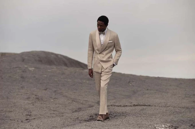Black man wearing a beige double-breasted suit walking on a barren dusty terrain under an overcast sky.