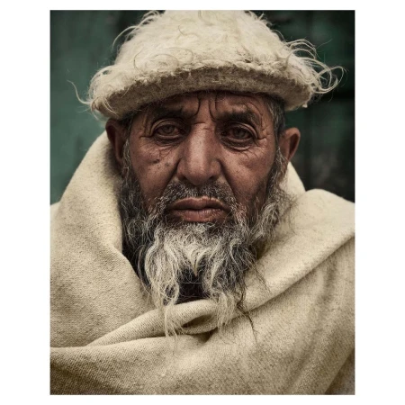 Close-up portrait of an elderly man with a long grey beard wearing a woolen cap and wrapped in a heavy shawl.