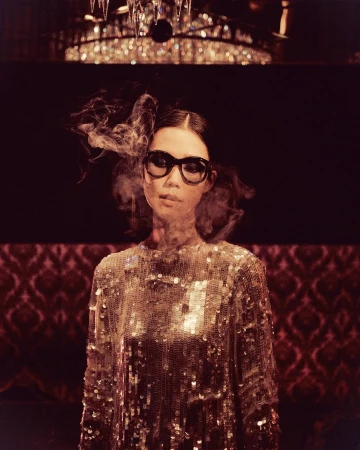 Woman wearing oversized black sunglasses and a sequined dress with wisps of smoke swirling around her head in a dimly lit interior.