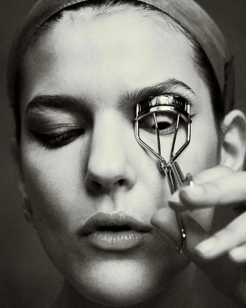 Close-up black and white portrait of a woman using an eyelash curler by photographer Alistair Taylor-Young.
