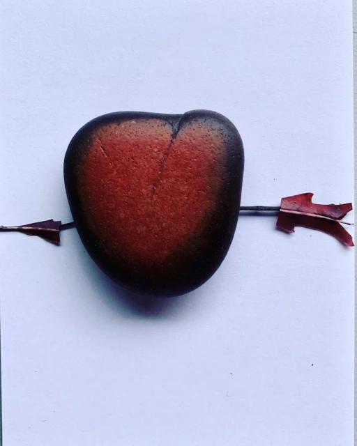 Heart-shaped stone with an arrow through it on a white surface. The arrow is made of twigs and red leaves, symbolizing love.