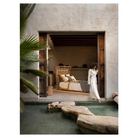 Woman in a robe steps across stones in a shallow pool outdoors leading to a bedroom with a rattan bed and palm tree.