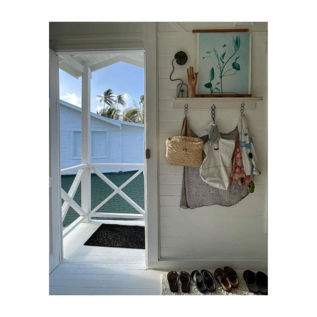 Bright, airy home interior with a view to a tropical exterior. A straw bag, clothing, and decorative art hang on a white wall.