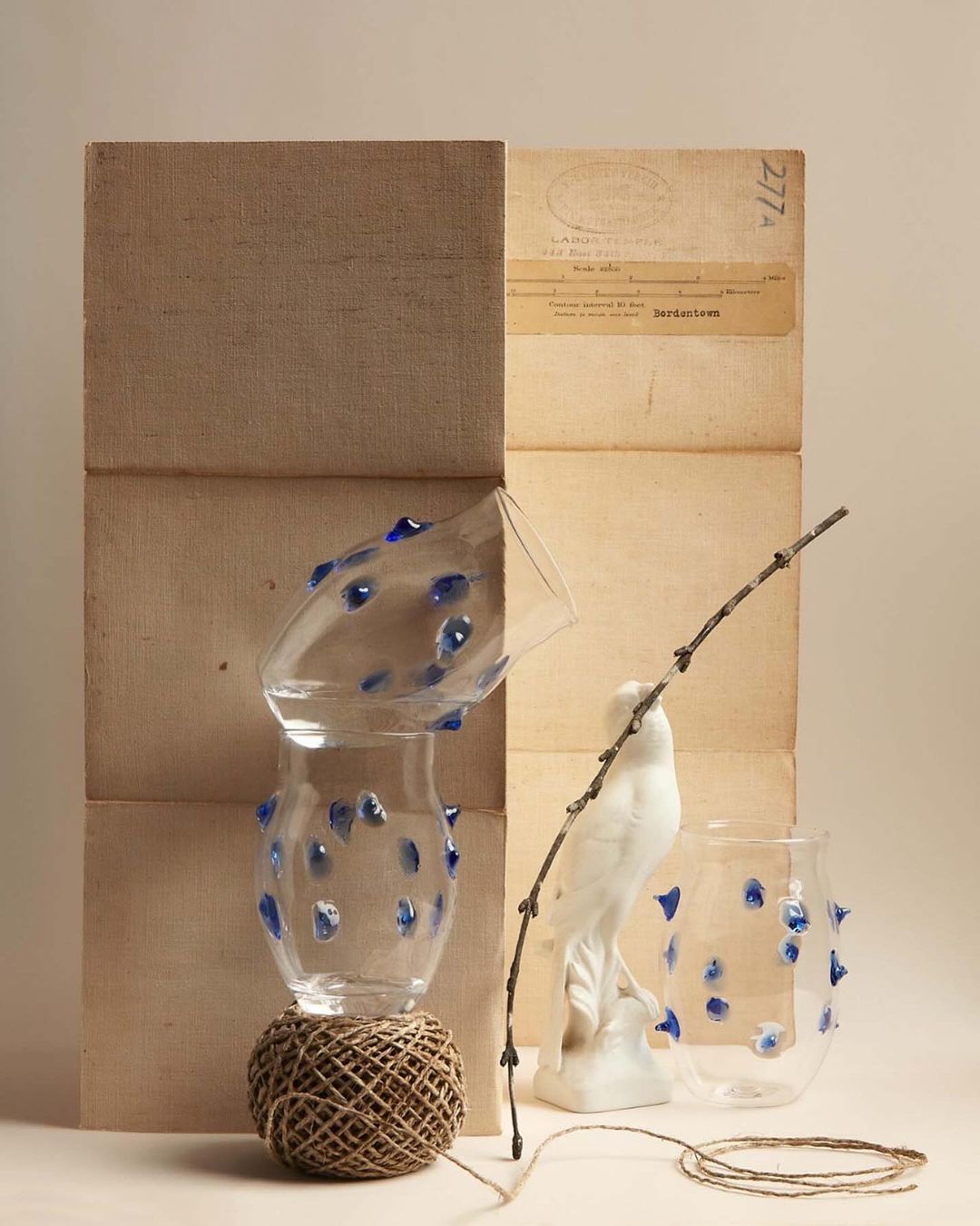 Still life composition featuring stacked vases adorned with blue accents, balanced on twine, alongside a porcelain bird and bare branch against a neutral backdrop.