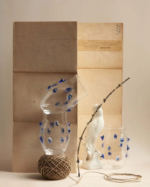 Still life composition featuring stacked vases adorned with blue accents, balanced on twine, alongside a porcelain bird and bare branch against a neutral backdrop.