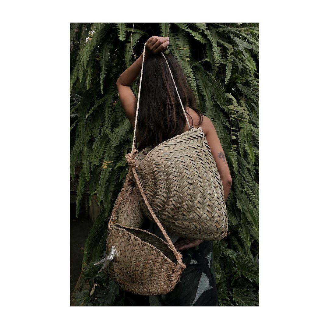 Woman with long hair carries two woven tote bags outdoors in front of lush green foliage in a natural light setting.