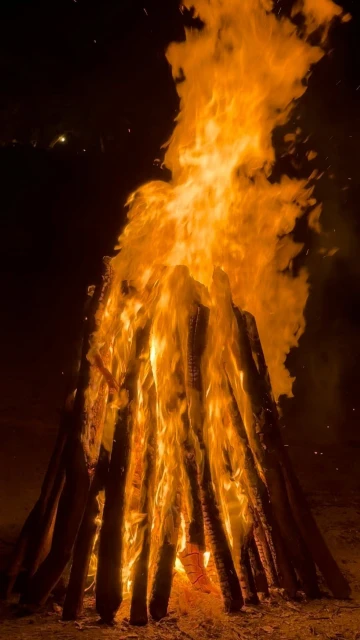 Large bonfire burns brightly at night, illuminating stacked logs with intense orange and yellow flames against a dark sky.