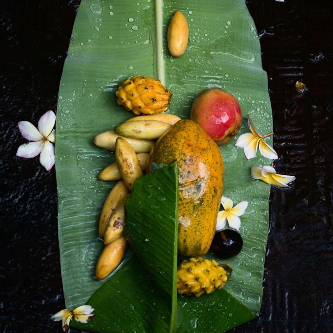 Overhead view of tropical fruits like papaya, mango, and dragon fruit arranged on a large green banana leaf, some white flowers.