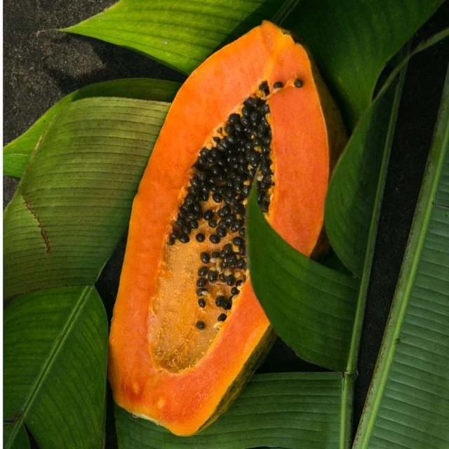 A halved papaya displays its orange flesh and black seeds, surrounded by large, green banana leaves on a textured surface.