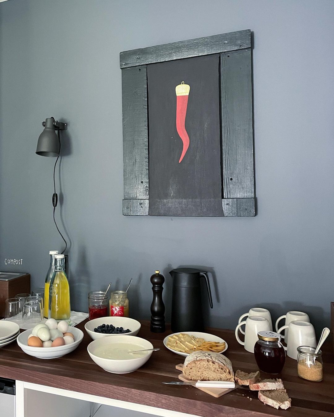 Breakfast buffet table with bread, fruit, eggs, juice, and jams against a grey wall with a framed chili pepper art print.