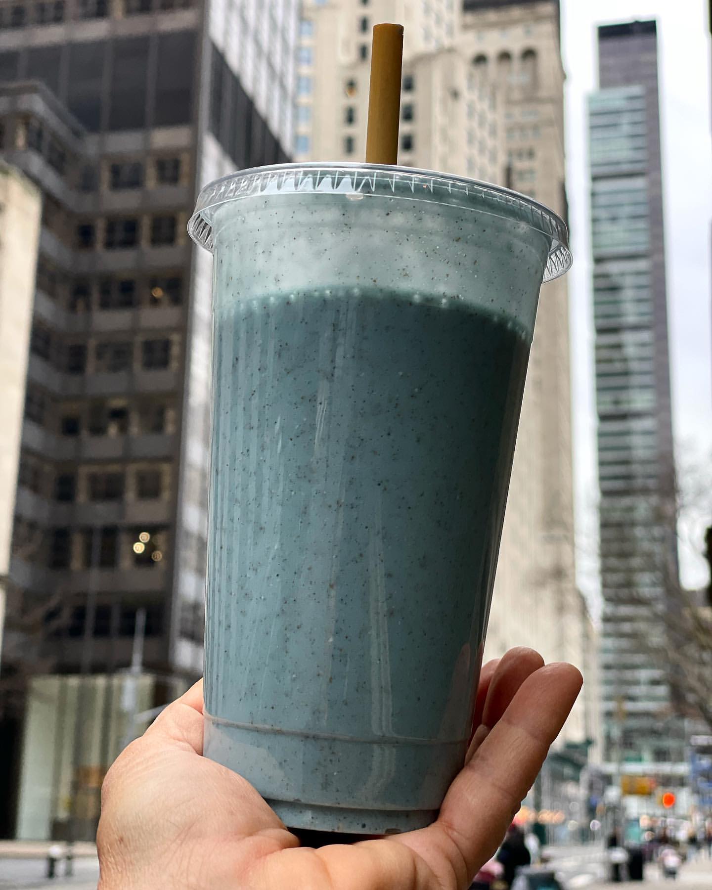 A clear plastic cup with a blue smoothie and a brown straw held in a hand, with tall city buildings blurred in the background.