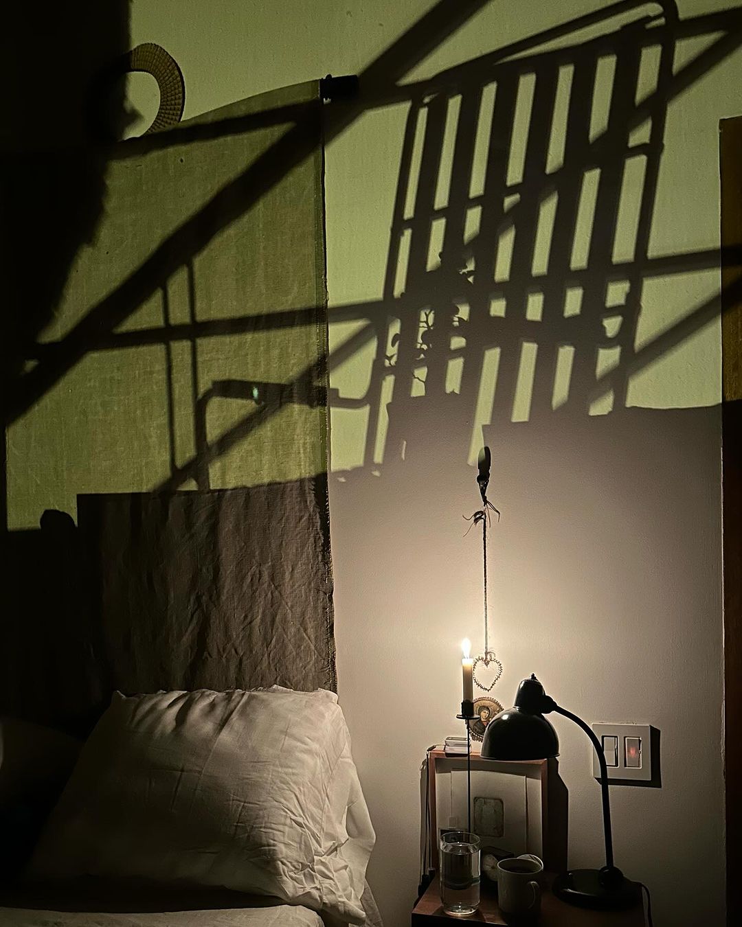 Bedroom interior with a bed, pillow, nightstand, lamp, candle, and shadows cast on the wall in soft, warm light.