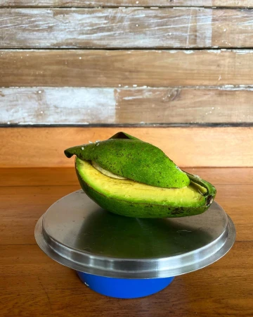 A halved avocado on a metal plate, showcasing its green skin, yellow flesh, and inner pit, sitting on a wooden surface with a plank background.