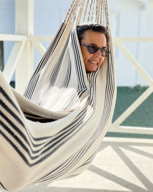 Adult woman lounges in a striped hammock on a porch. She wears sunglasses and smiles in the sunlight with white siding in background.