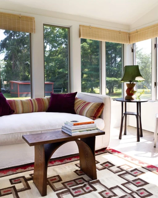 Bright sunroom with a cream sofa topped with pillows, a wooden table, patterned rug, and view of trees and a shed through the windows.