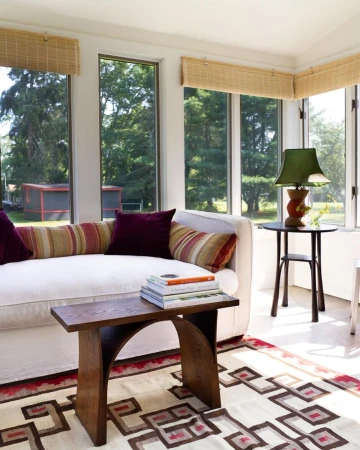 Bright sunroom with a cream sofa topped with pillows, a wooden table, patterned rug, and view of trees and a shed through the windows.
