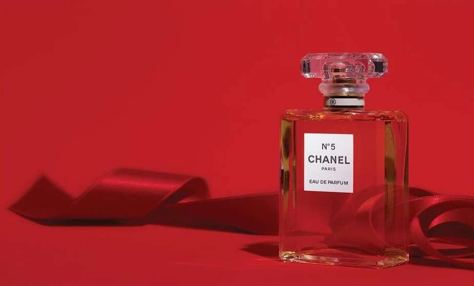 Chanel N°5 perfume bottle on a red background with a red satin ribbon, featuring a rectangular glass bottle and crystal-like stopper.