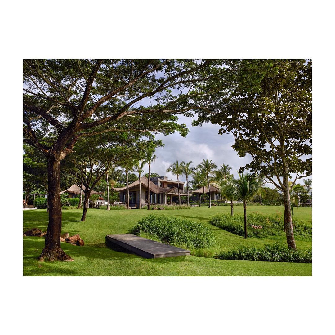 Exterior view of a large, modern home with a straw roof surrounded by palm trees and green grass, showcasing architectural design and landscaping.