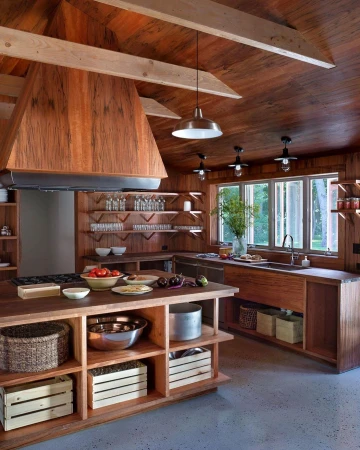 Warm, wood-clad kitchen interior with island, open shelving, fruits, vegetables, bowls, cookware, hanging lights, and outdoor views through windows.
