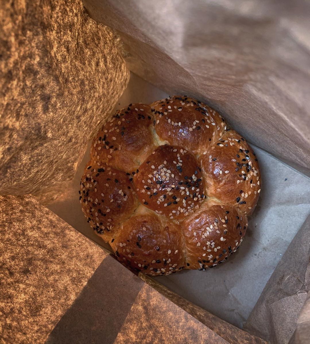 Overhead shot of golden brown challah bread with sesame seeds inside a brown paper bag, showcasing its braided texture and shape.