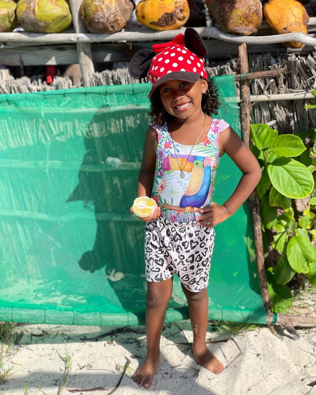 A smiling young girl in a colorful swimsuit and Minnie Mouse hat holds a lemon slice on a sandy beach with coconuts overhead.