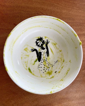 White ceramic bowl with green food residue and an interior black illustration of a figure, on a wooden table.