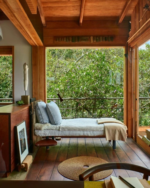 Indoor reading nook featuring a daybed, artwork, and a view of lush greenery through a large window in a wood-paneled room.