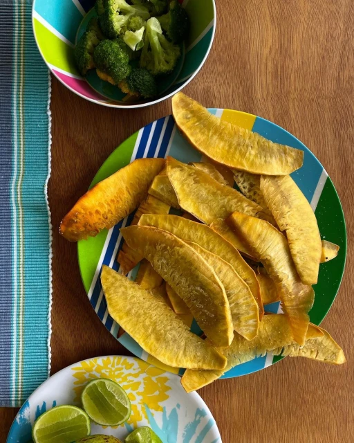 Overhead view of golden fried plantain slices on a striped plate, accompanied by broccoli and halved limes, all arranged on a wooden surface.