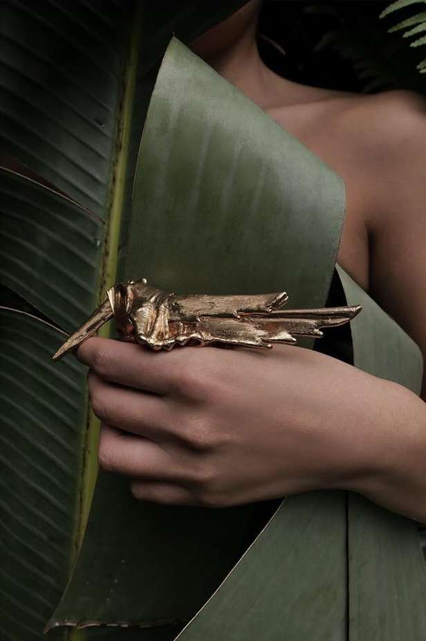 Close-up of a hand displaying a large, golden insect-shaped ring against a backdrop of lush green banana leaves, showcasing nature-inspired jewelry.